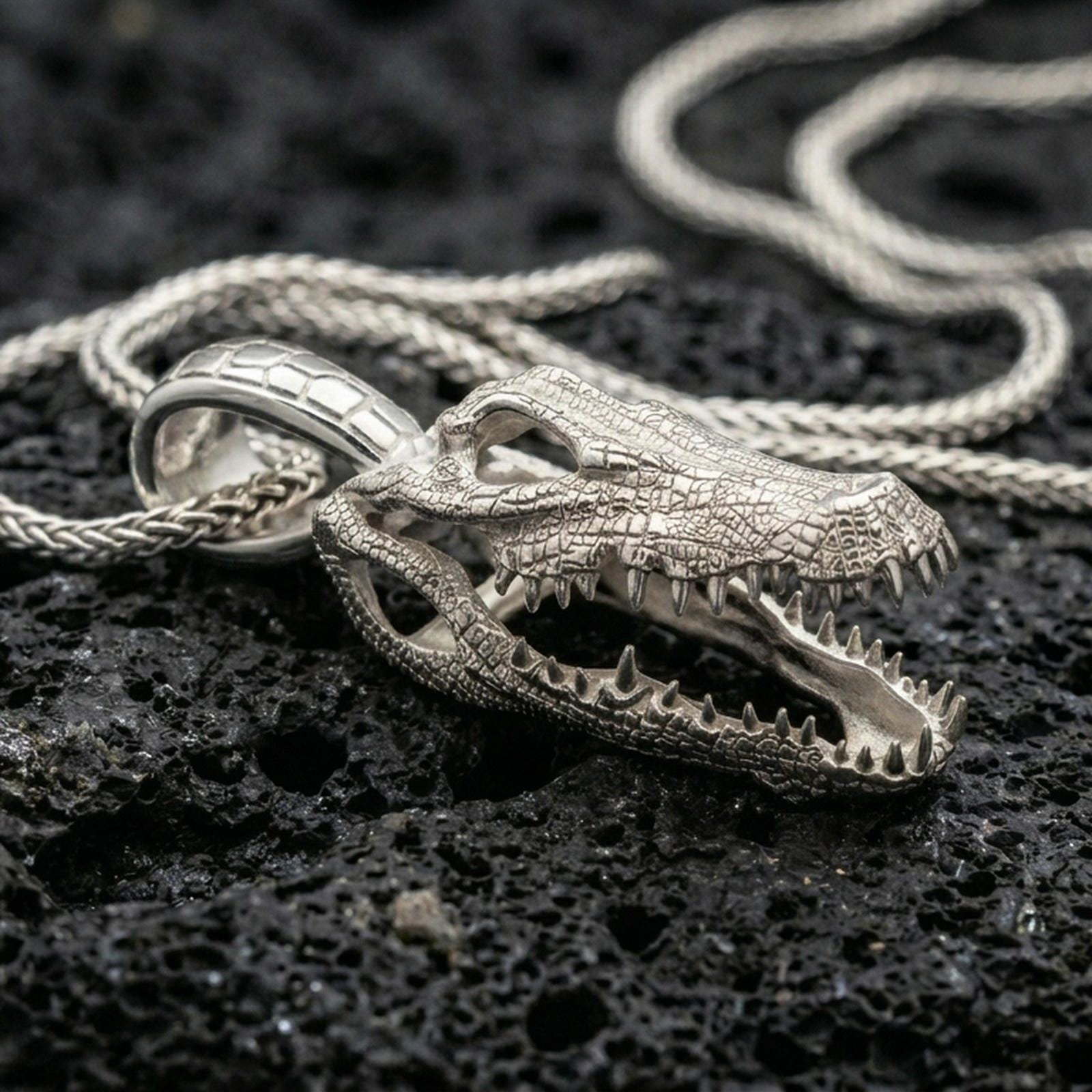 Polished silver gator skull necklace with sculpted reptile head, sharp teeth detailing, and realistic skull structure placed on textured volcanic rock in a premium studio product shot