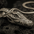 Load image into Gallery viewer, Bronze alligator skull pendant with open jaw, detailed reptile bone texture, and fierce animal silhouette displayed on dark volcanic rock in a close-up luxury jewelry composition
