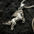 Load image into Gallery viewer, Polished silver horse necklace with sculpted rearing stallion design, dynamic body contours, and elegant flowing tail placed on textured volcanic rock in a premium studio product shot
