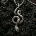 Load image into Gallery viewer, Oxidized snake pendant with coiled serpent silhouette and detailed scale texture displayed on dark volcanic rock in a close-up luxury jewelry composition
