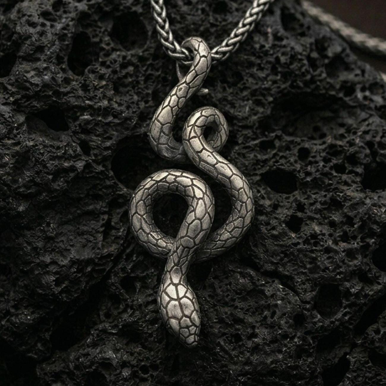 Oxidized snake pendant with coiled serpent silhouette and detailed scale texture displayed on dark volcanic rock in a close-up luxury jewelry composition