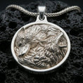 Load image into Gallery viewer, Polished silver wolf necklace with detailed twin wolf design, sculpted fur texture, and circular wilderness-inspired frame placed on textured volcanic rock in a premium studio product shot
