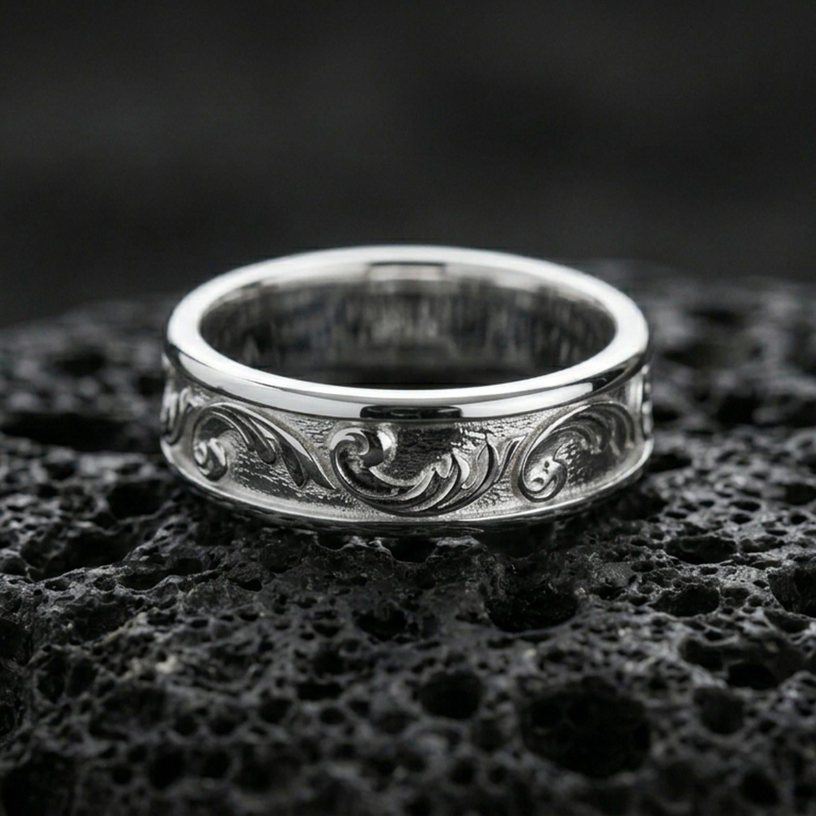 Polished silver floral engraved wedding band on volcanic rock, bright smooth edges with crisp scrolling leaf motifs in macro detail.