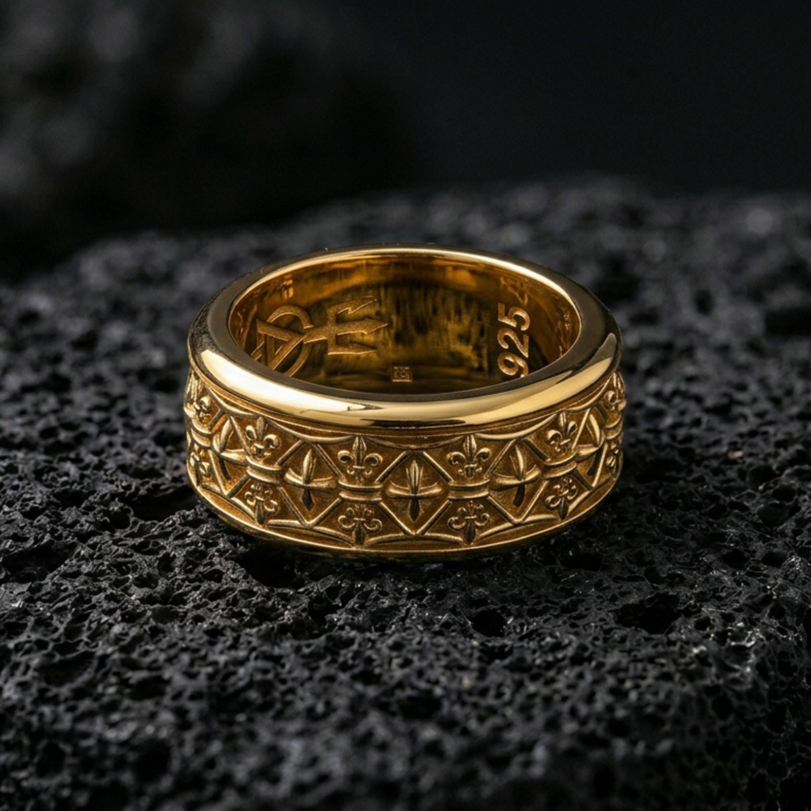 Fleur de lis patterned ring displayed on volcanic rock with a gold plated finish, highlighting the raised symbols and textured band.