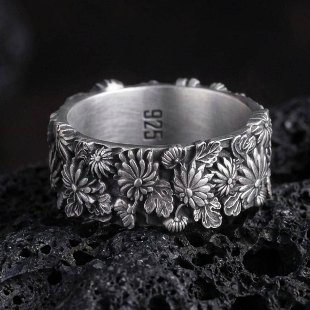 Sterling silver floral band ring photographed on volcanic rock with oxidized contrast highlighting the raised daisy and leaf relief.