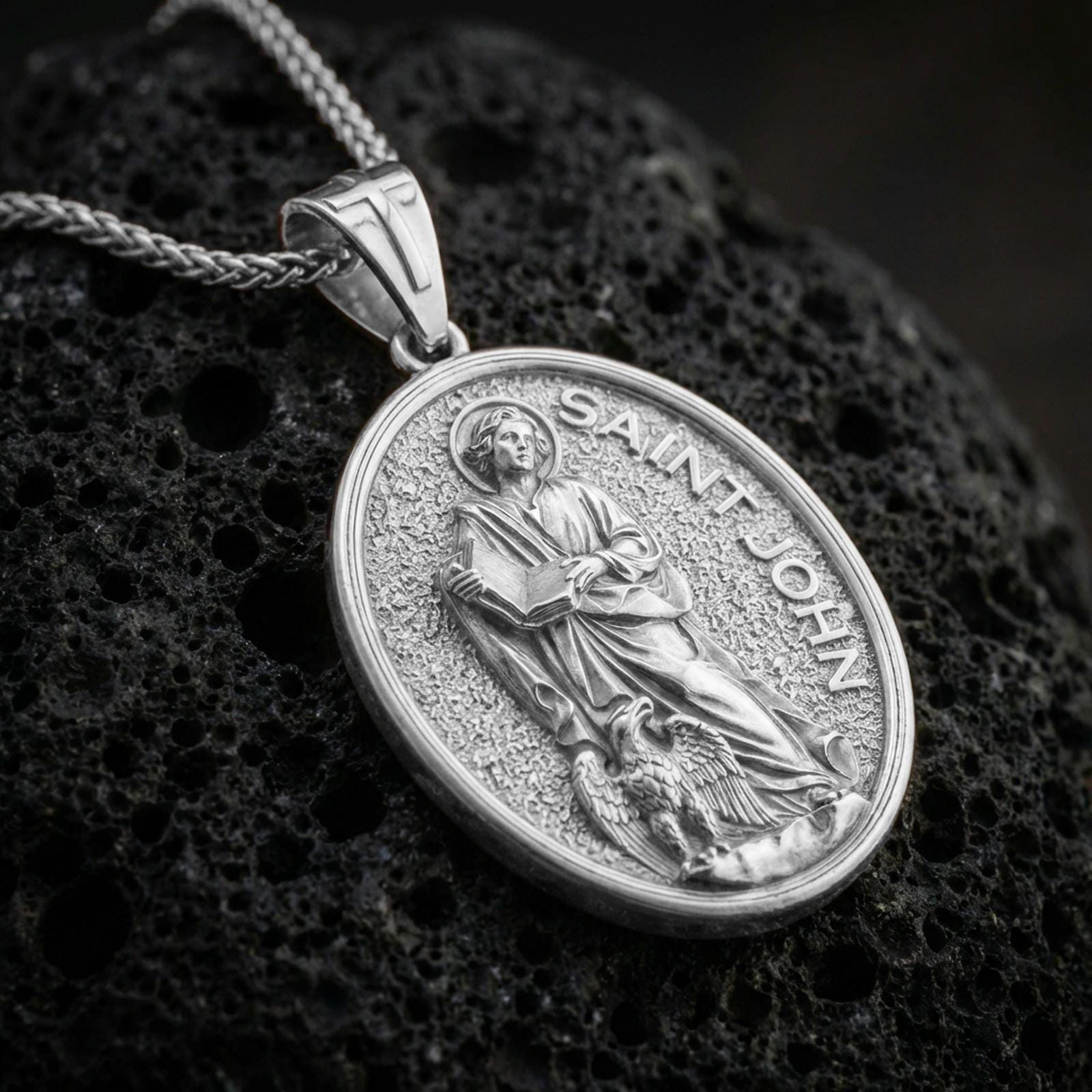 A polished 925 sterling silver Saint John the Evangelist pendant on volcanic rock, oval saint medal with detailed relief.