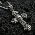 Load image into Gallery viewer, Oxidized Chicano tattoo style cross pendant with rose details and masked face motif displayed on dark volcanic rock in a close-up luxury jewelry shot
