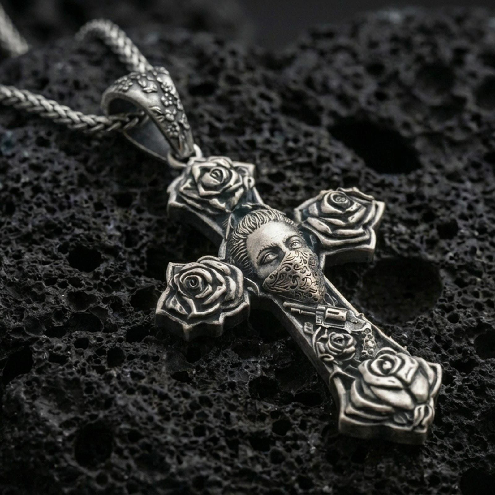 Oxidized Chicano tattoo style cross pendant with rose details and masked face motif displayed on dark volcanic rock in a close-up luxury jewelry shot