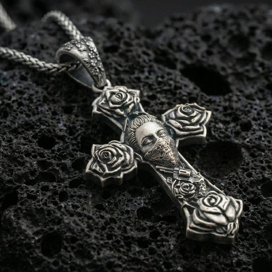 Oxidized Chicano tattoo style cross pendant with rose details and masked face motif displayed on dark volcanic rock in a close-up luxury jewelry shot