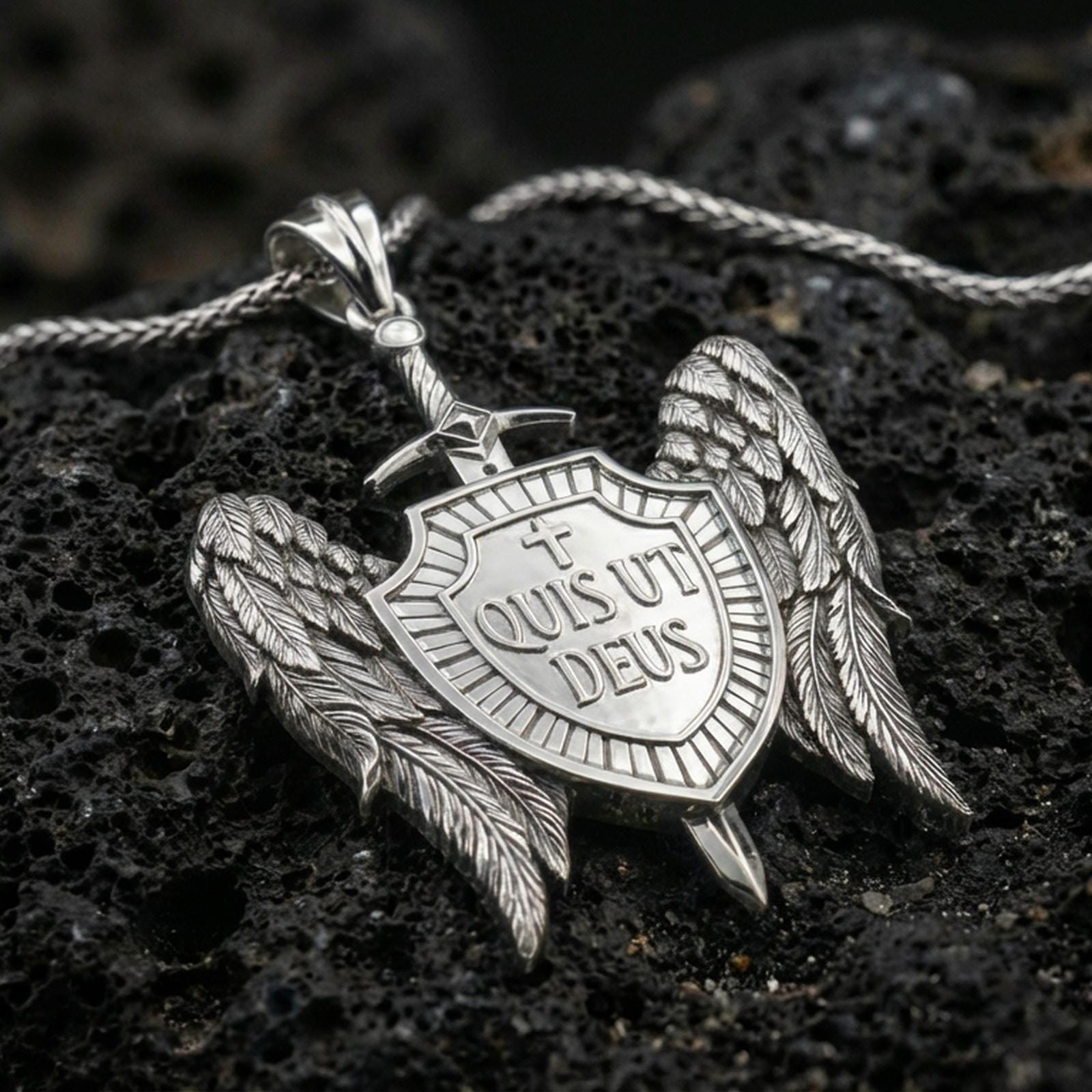 Polished silver spiritual protection pendant with winged shield design and Quis Ut Deus text placed on textured volcanic rock in a premium studio composition
