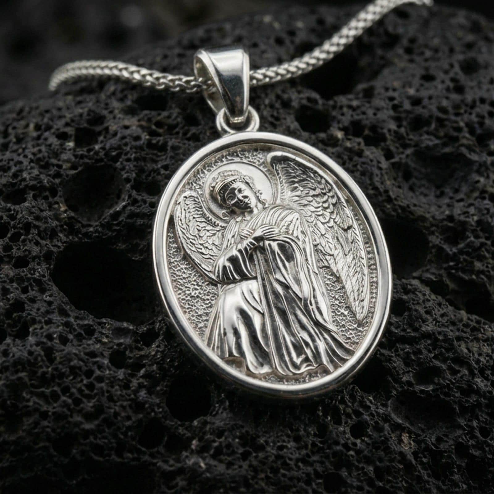 Polished silver religious medal pendant with a seated angel relief and oval border placed on textured volcanic rock in a premium studio product shot