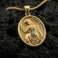 Load image into Gallery viewer, Gold plated angel medallion pendant with sculpted wings and devotional figure showcased on black volcanic rock in a refined macro jewelry image

