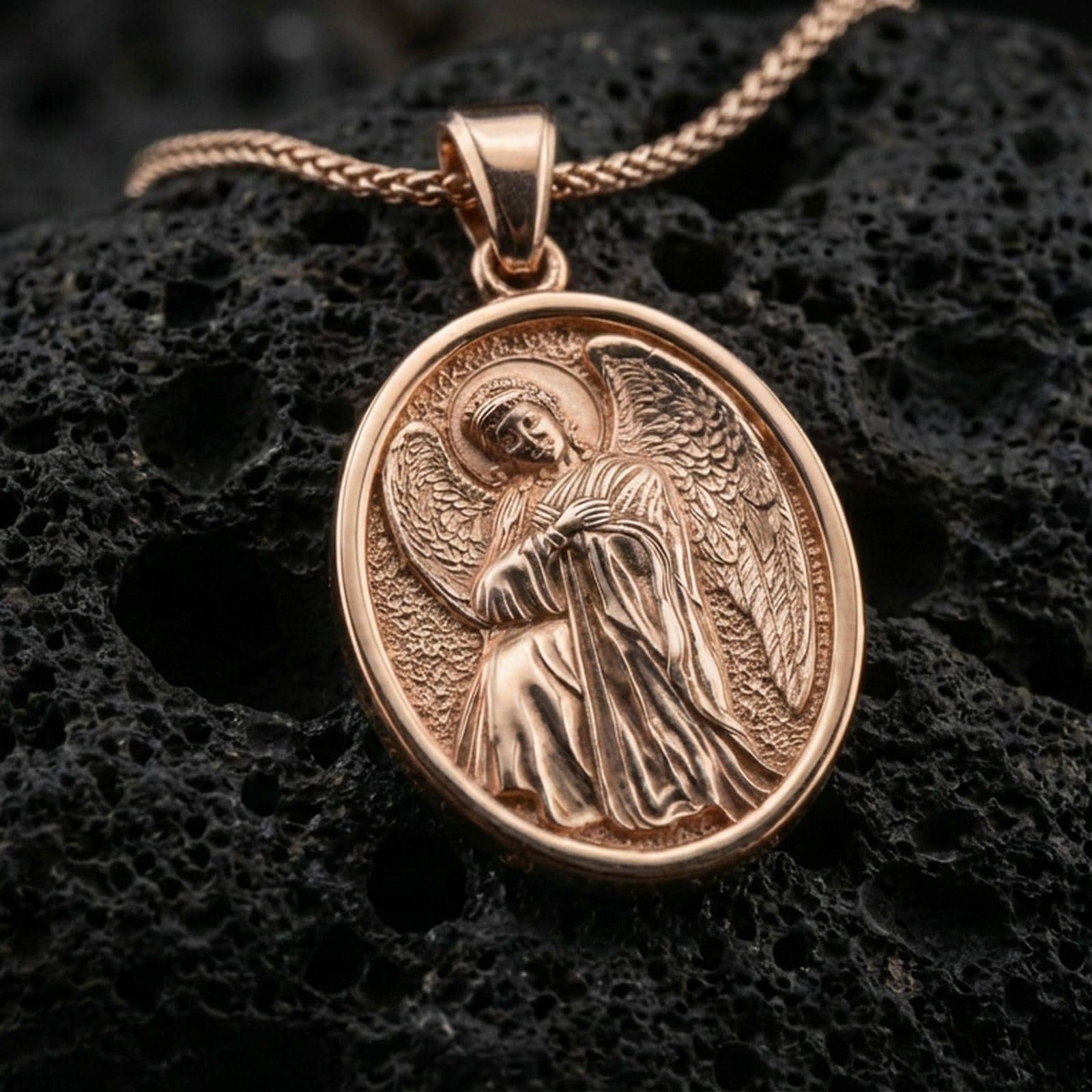 Rose gold plated oval angel pendant with detailed halo, robes, and wing relief resting on volcanic rock in an elegant close-up product presentation