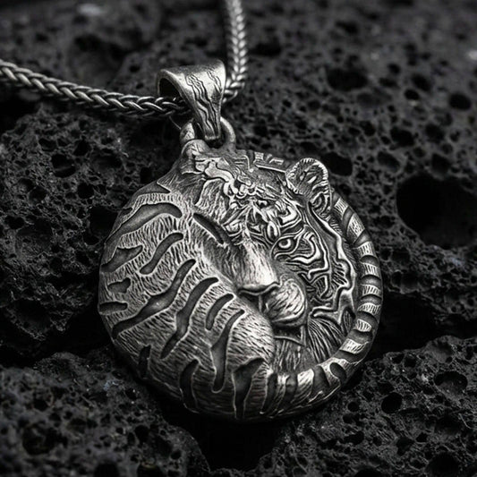 Oxidized tiger pendant with curled sleeping feline form, detailed stripe engraving, and round symbolic jungle silhouette displayed on dark volcanic rock in a close-up luxury jewelry composition