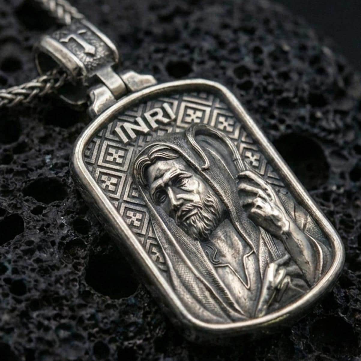 Oxidized Jesus pendant with INRI inscription and shepherd staff relief displayed on dark volcanic rock in a close-up luxury jewelry composition