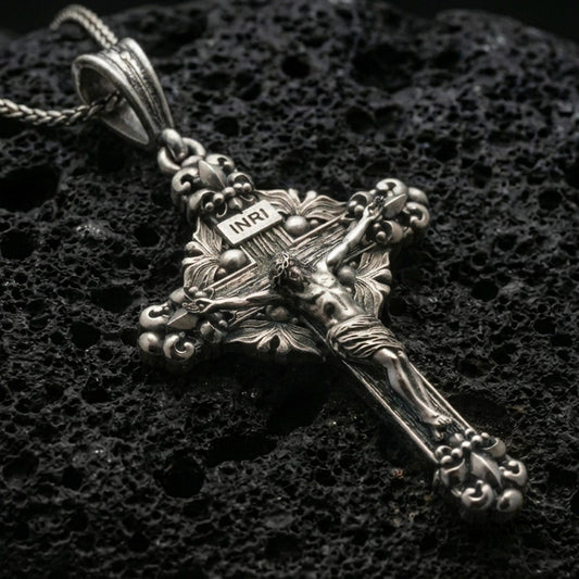 Oxidized crucifix pendant with INRI plaque, detailed Jesus figure, and ornate baroque cross design displayed on dark volcanic rock in a close-up luxury jewelry composition