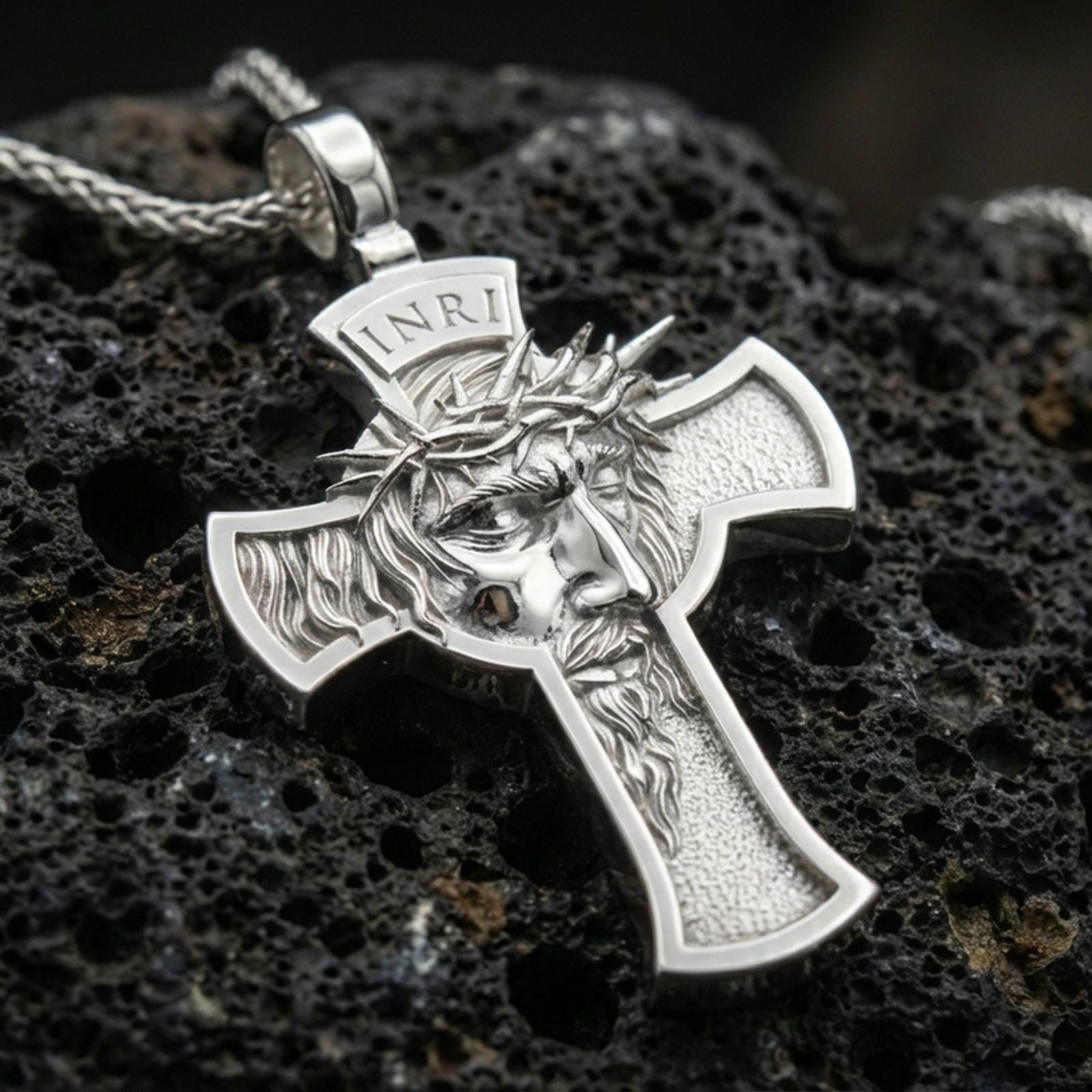 Polished silver Christian cross pendant with detailed Jesus portrait, crown of thorns, and open cross silhouette placed on textured volcanic rock in a premium studio product shot