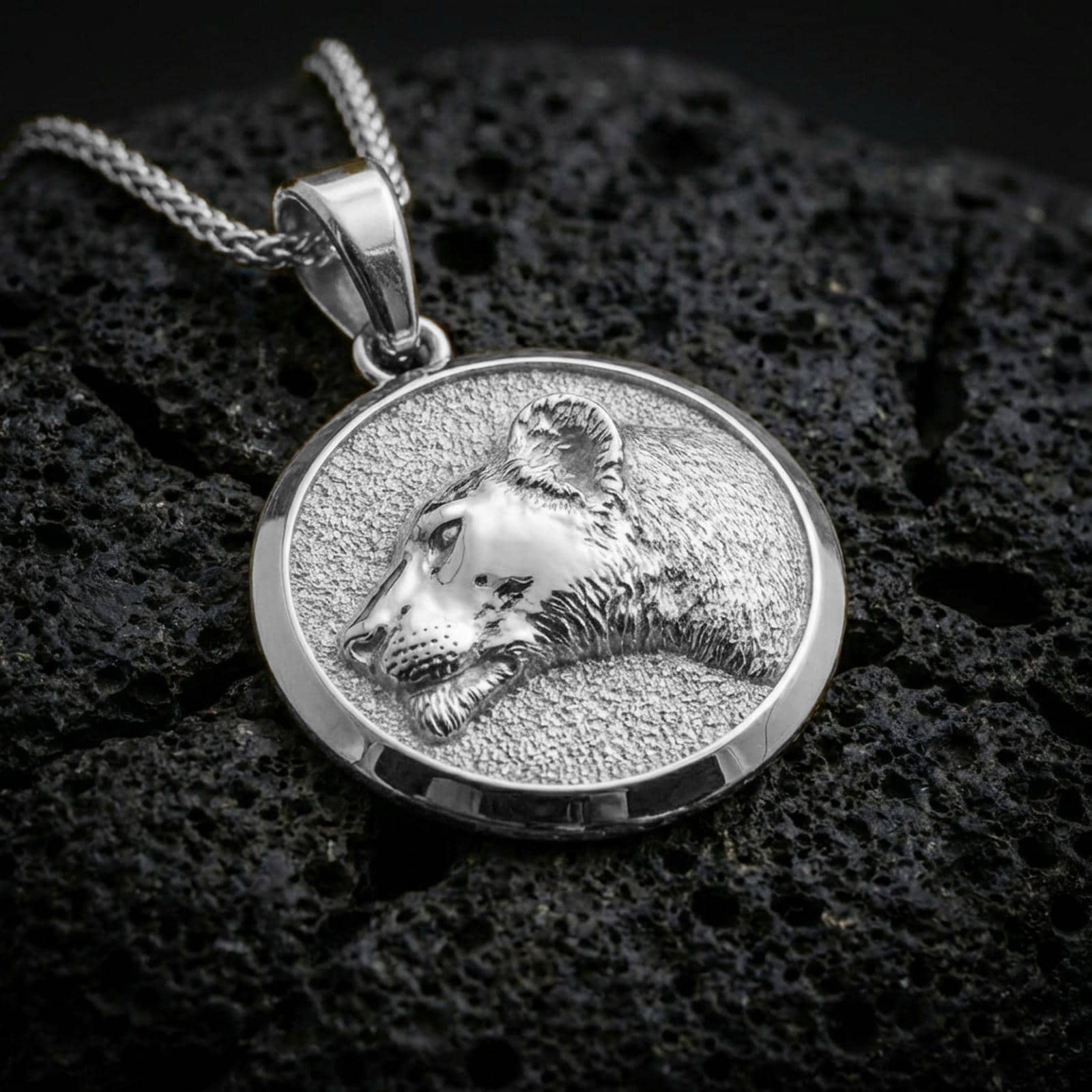 Polished silver lioness necklace with sculpted female lion portrait, textured fur detailing, and refined circular medallion design placed on textured volcanic rock in a premium studio product shot