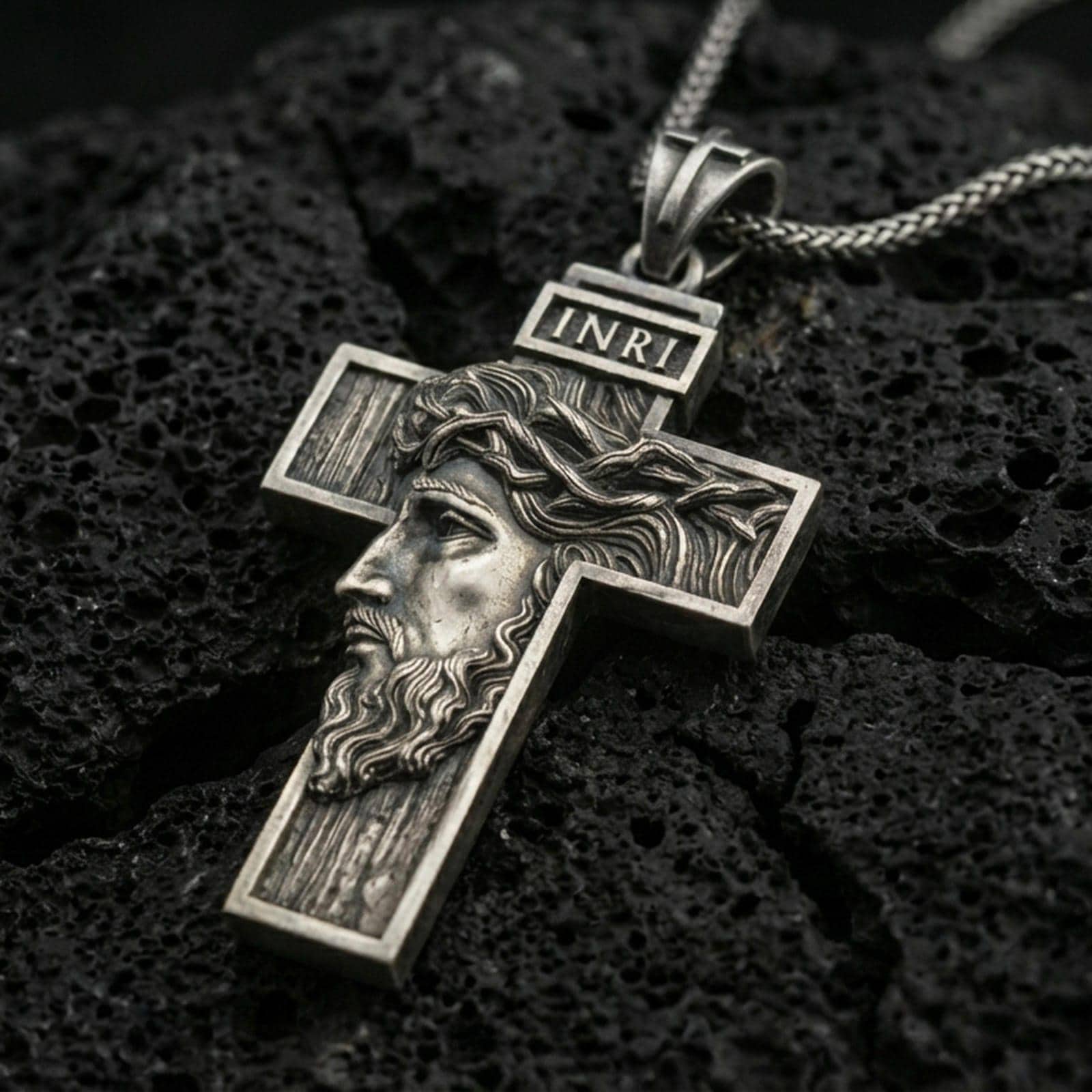 Oxidized Jesus cross pendant with side profile portrait, crown of thorns, and INRI detail displayed on dark volcanic rock in a close-up luxury jewelry composition