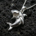 Load image into Gallery viewer, Polished silver shark necklace with detailed teeth, dynamic swimming pose, and bold ocean-inspired animal form placed on textured volcanic rock in a premium studio product shot
