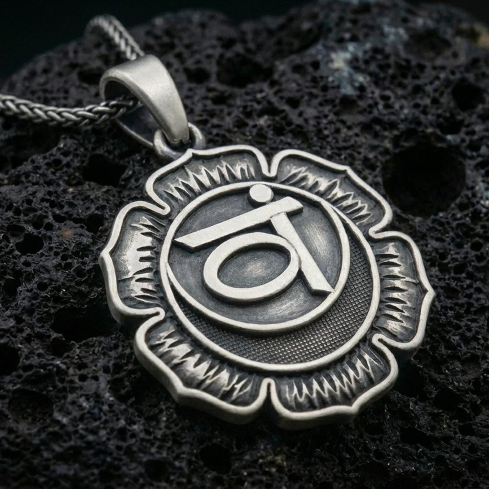 Oxidized throat chakra pendant with lotus flower silhouette and sacred symbol relief displayed on dark volcanic rock in a close-up luxury jewelry composition