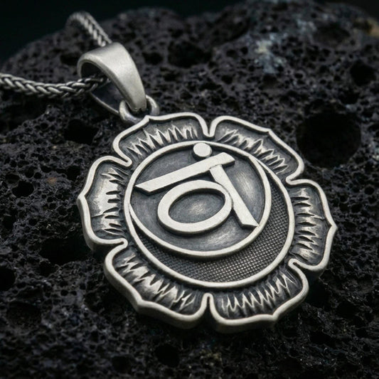 Oxidized throat chakra pendant with lotus flower silhouette and sacred symbol relief displayed on dark volcanic rock in a close-up luxury jewelry composition