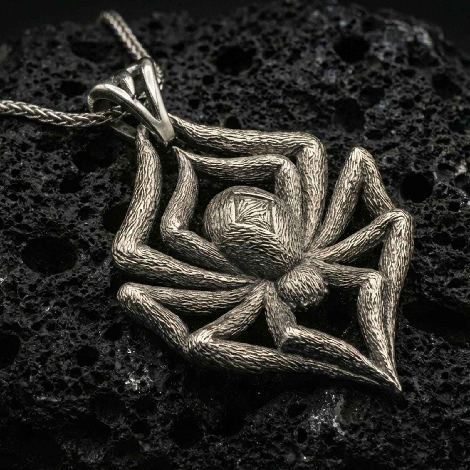 Oxidized spider pendant with textured legs, sculpted body relief, and bold arachnid silhouette displayed on dark volcanic rock in a close-up luxury jewelry composition
