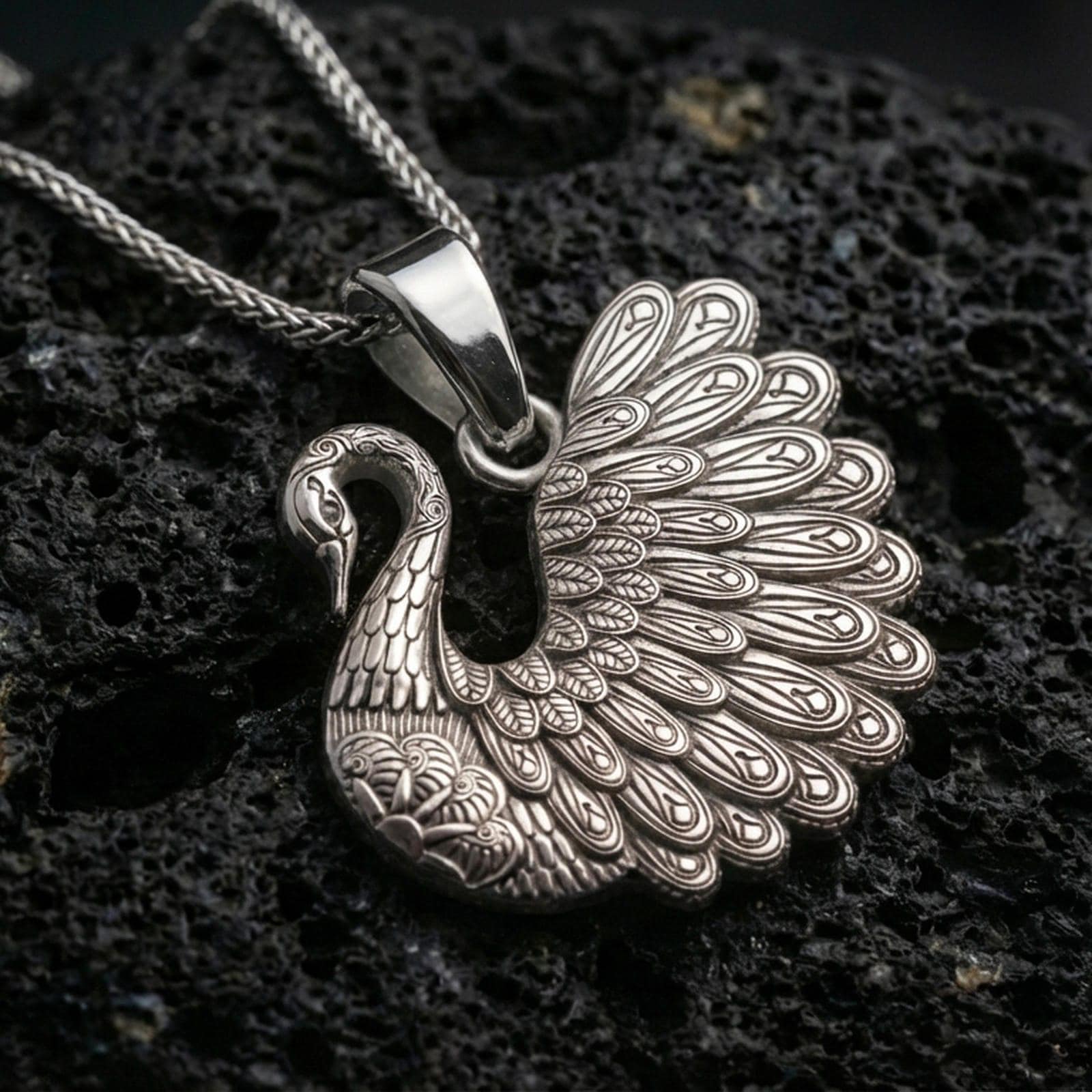 Polished silver swan necklace with layered feather detailing, sculpted wing fan design, and refined bird motif placed on textured volcanic rock in a premium studio product shot