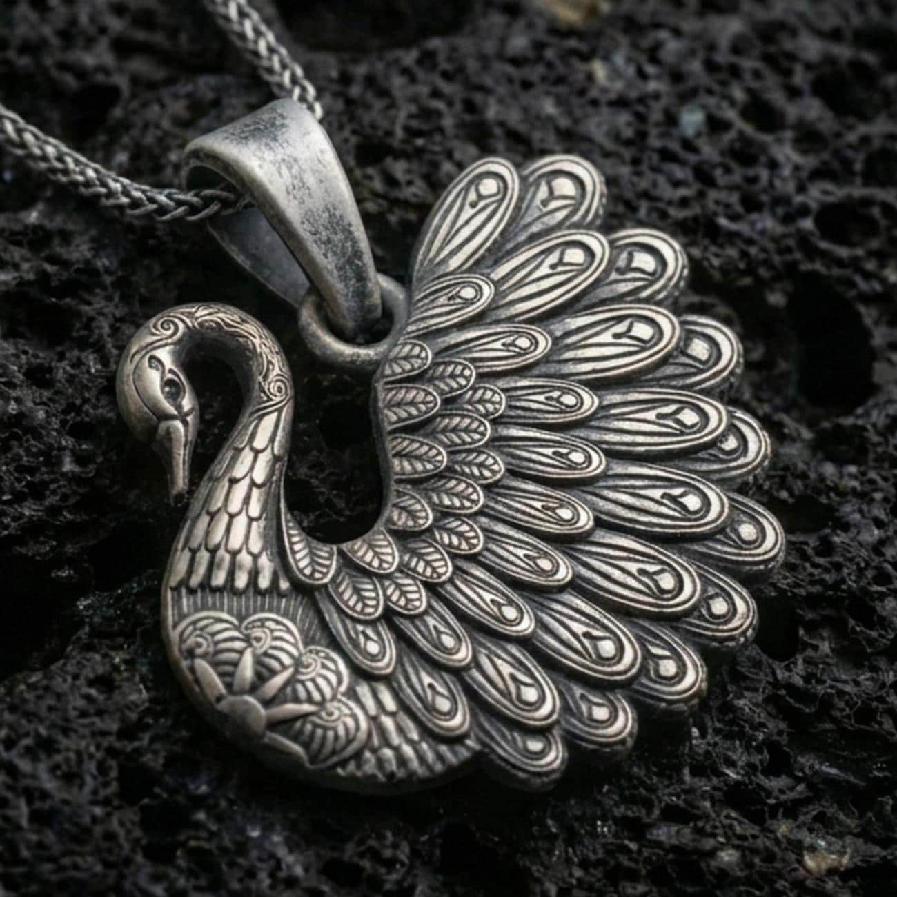 Oxidized swan pendant with detailed feather engraving, graceful curved neck, and elegant bird silhouette displayed on dark volcanic rock in a close-up luxury jewelry composition