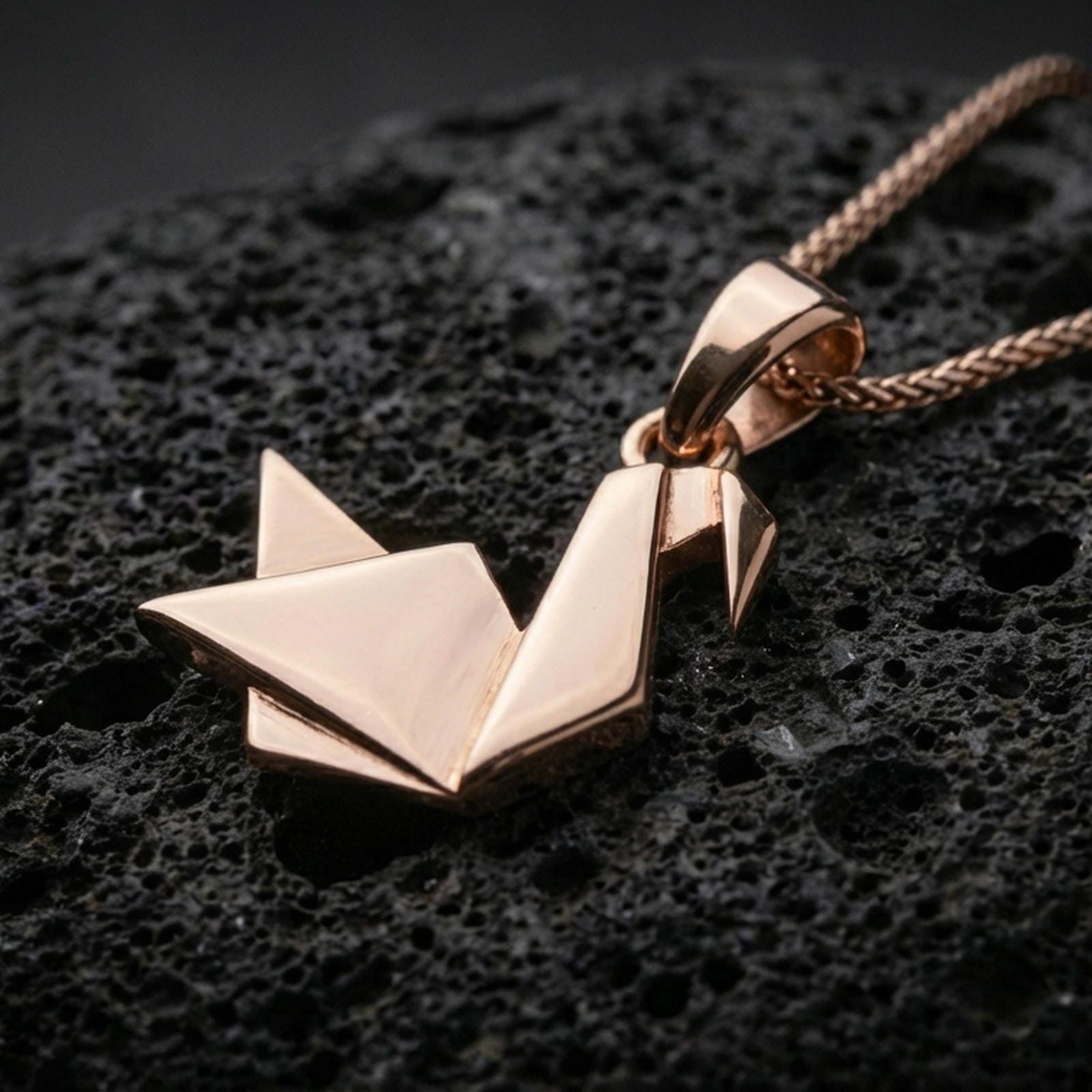 Rose gold plated geometric swan necklace with folded origami-inspired form, sharp angular planes, and elegant minimalist bird design resting on volcanic rock in an elegant close-up product presentation