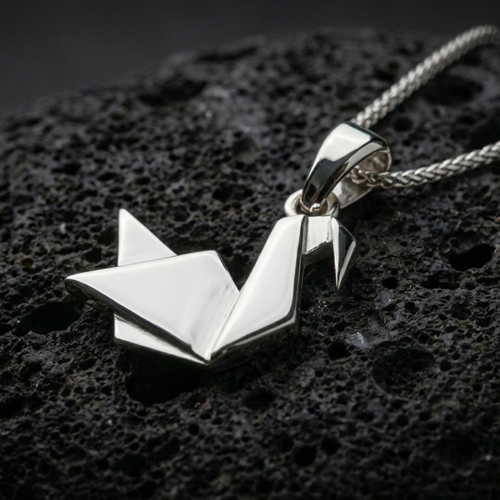Polished silver origami swan necklace with sharp folded contours, abstract geometric bird form, and minimalist sculptural design placed on textured volcanic rock in a premium studio product shot