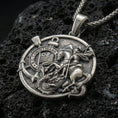 Load image into Gallery viewer, Oxidized Saint George pendant with dragon slayer scene, Corinthians inscription, and round medallion design displayed on dark volcanic rock in a close-up luxury jewelry composition
