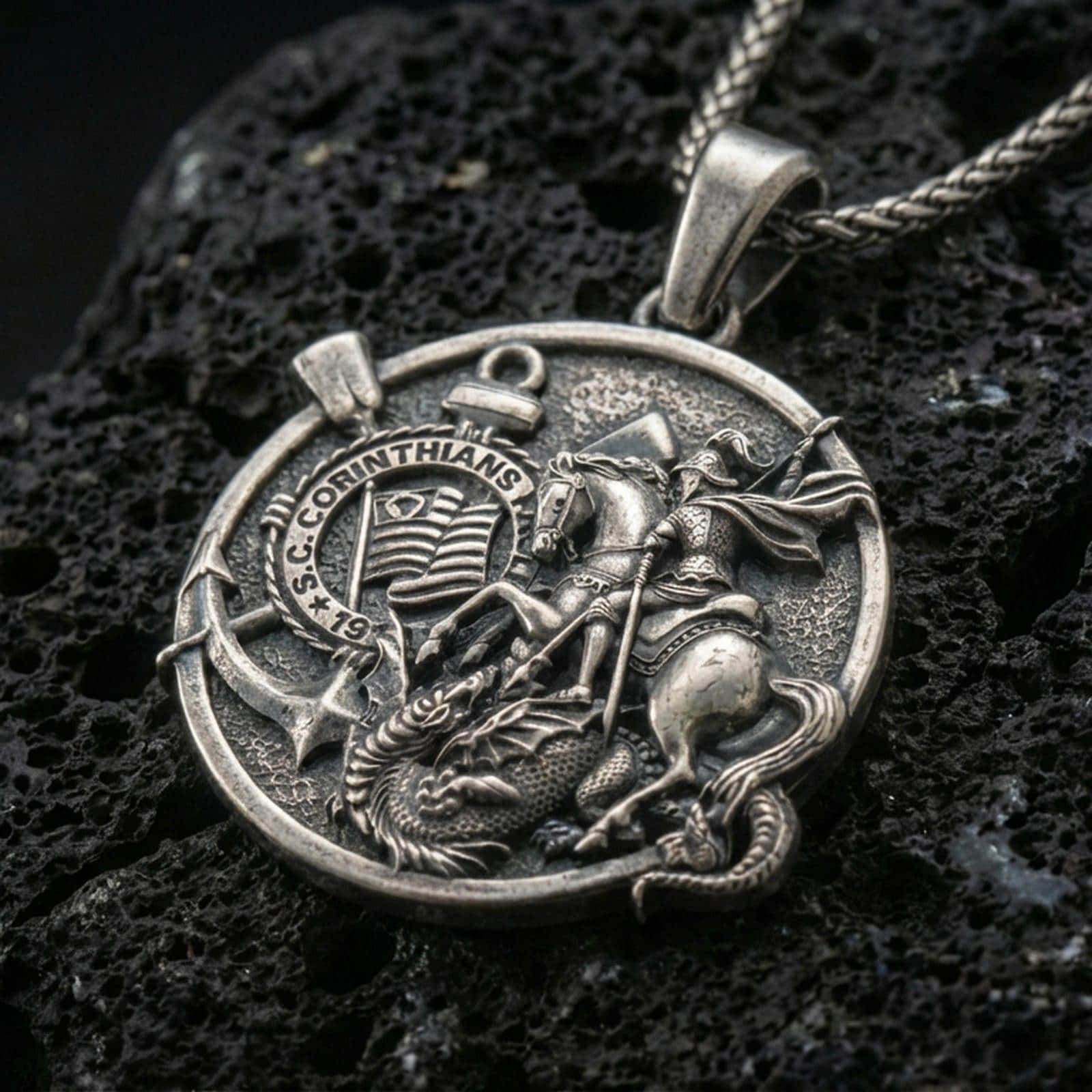 Oxidized Saint George pendant with dragon slayer scene, Corinthians inscription, and round medallion design displayed on dark volcanic rock in a close-up luxury jewelry composition