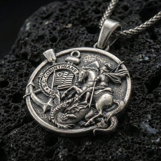 Oxidized Saint George pendant with dragon slayer scene, Corinthians inscription, and round medallion design displayed on dark volcanic rock in a close-up luxury jewelry composition