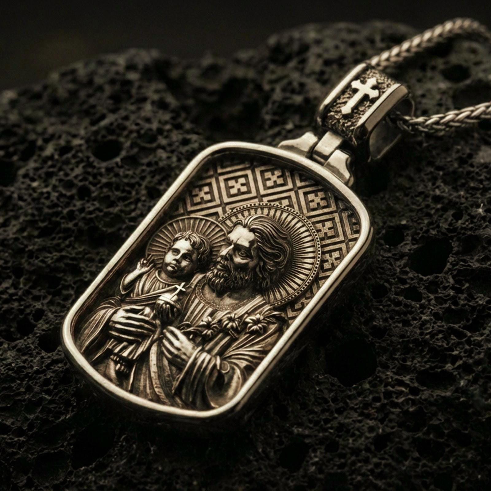 Bronze Saint Joseph pendant with Child Jesus, halo details, and framed devotional portrait displayed on dark volcanic rock in a close-up luxury jewelry composition