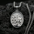 Load image into Gallery viewer, Polished silver religious oval pendant with a robed saint, lion motif, and engraved book placed on textured volcanic rock in a premium studio product shot
