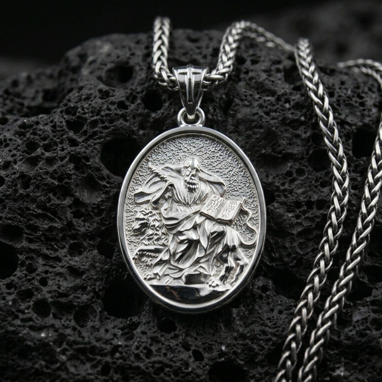 Polished silver religious oval pendant with a robed saint, lion motif, and engraved book placed on textured volcanic rock in a premium studio product shot