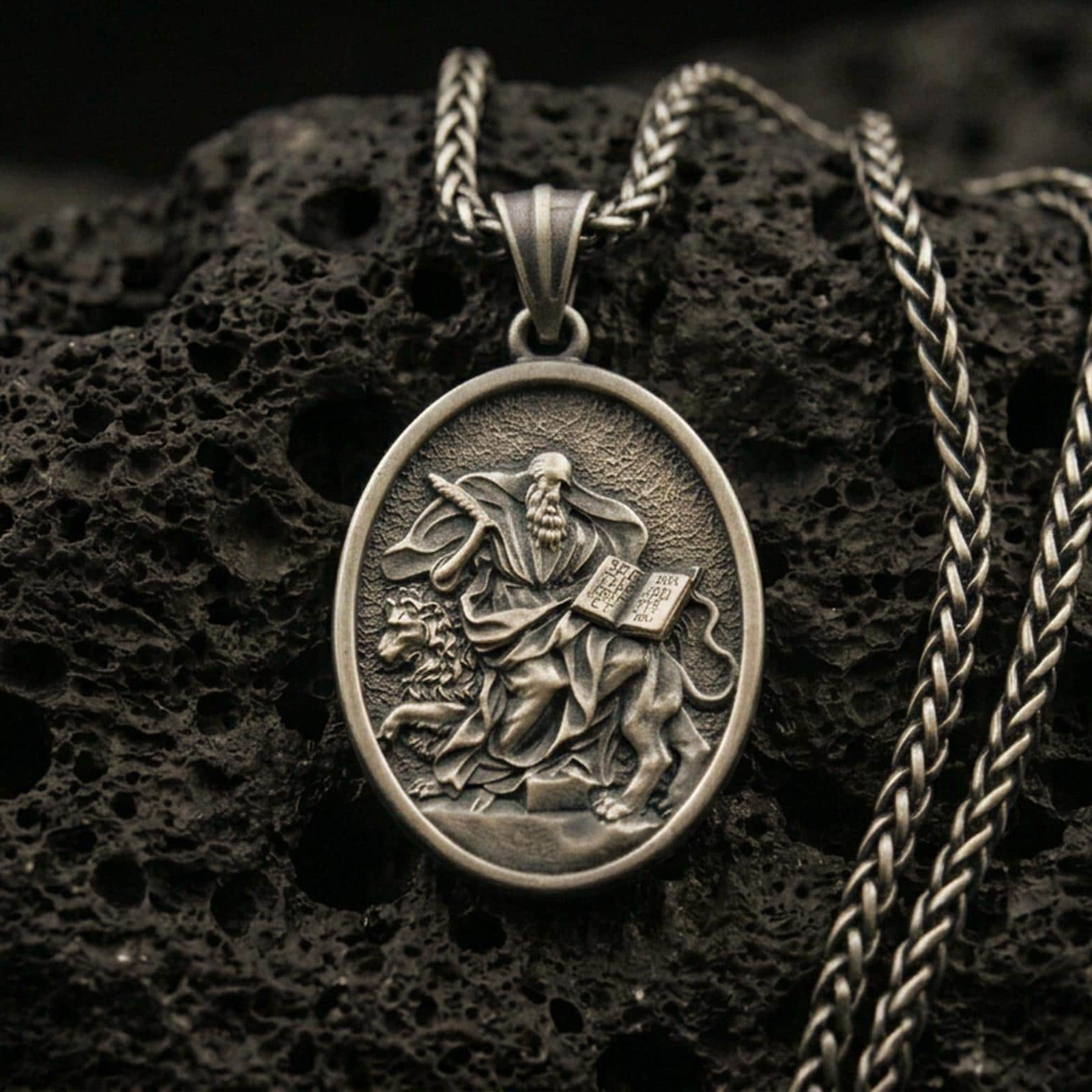 Bronze saint medallion pendant with a seated bearded figure, lion detail, and open scripture relief displayed on dark volcanic rock in a close-up luxury jewelry composition