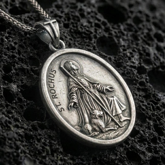Oxidized Saint Rochus medallion with pilgrim staff, halo, and dog companion displayed on dark volcanic rock in a close-up luxury jewelry composition