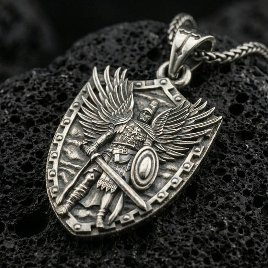 Oxidized Archangel Michael pendant with shield silhouette, winged warrior figure, sword, and armor detail displayed on dark volcanic rock in a close-up luxury jewelry composition