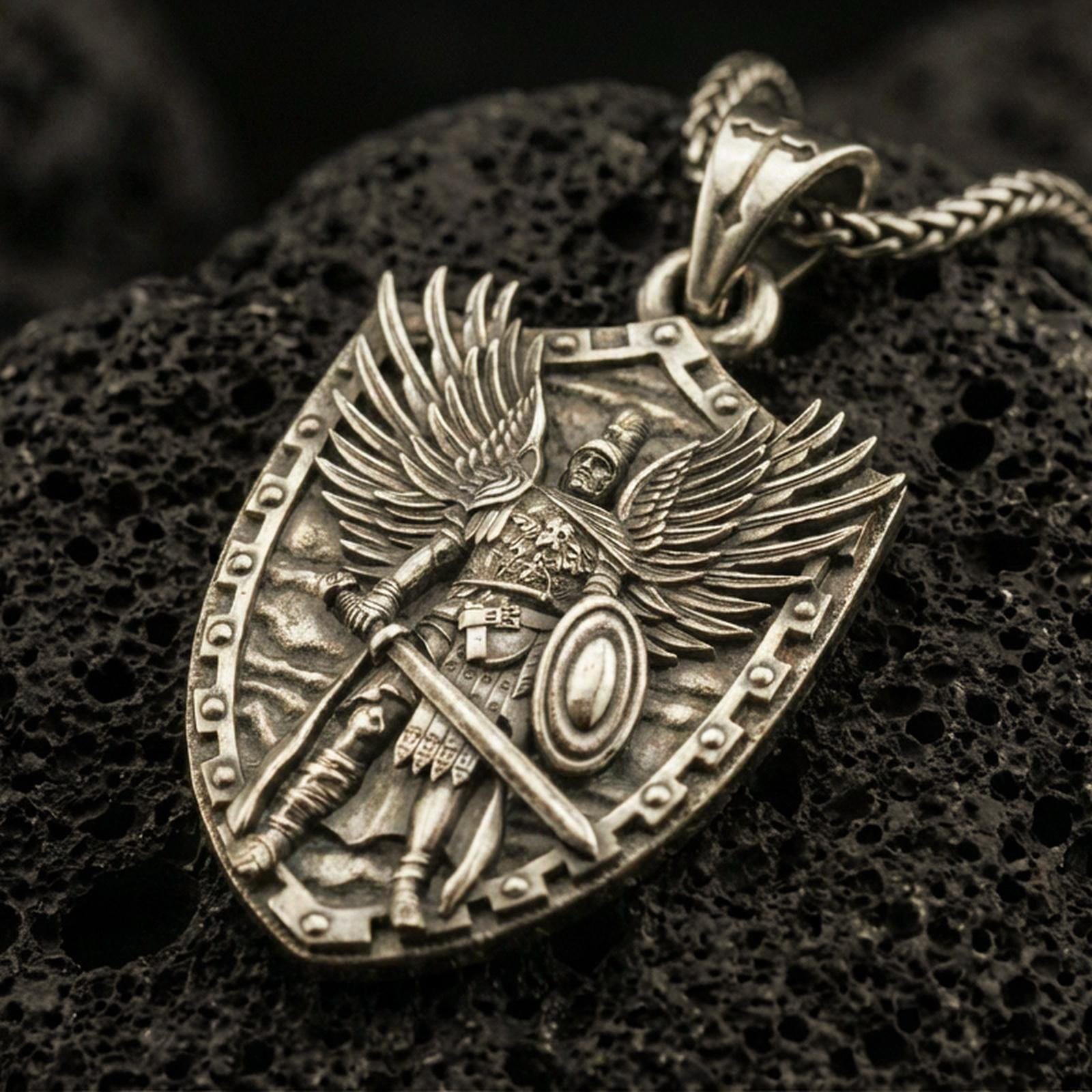 Bronze Archangel Michael pendant with shield silhouette, winged warrior figure, sword, and armor detail displayed on dark volcanic rock in a close-up luxury jewelry composition