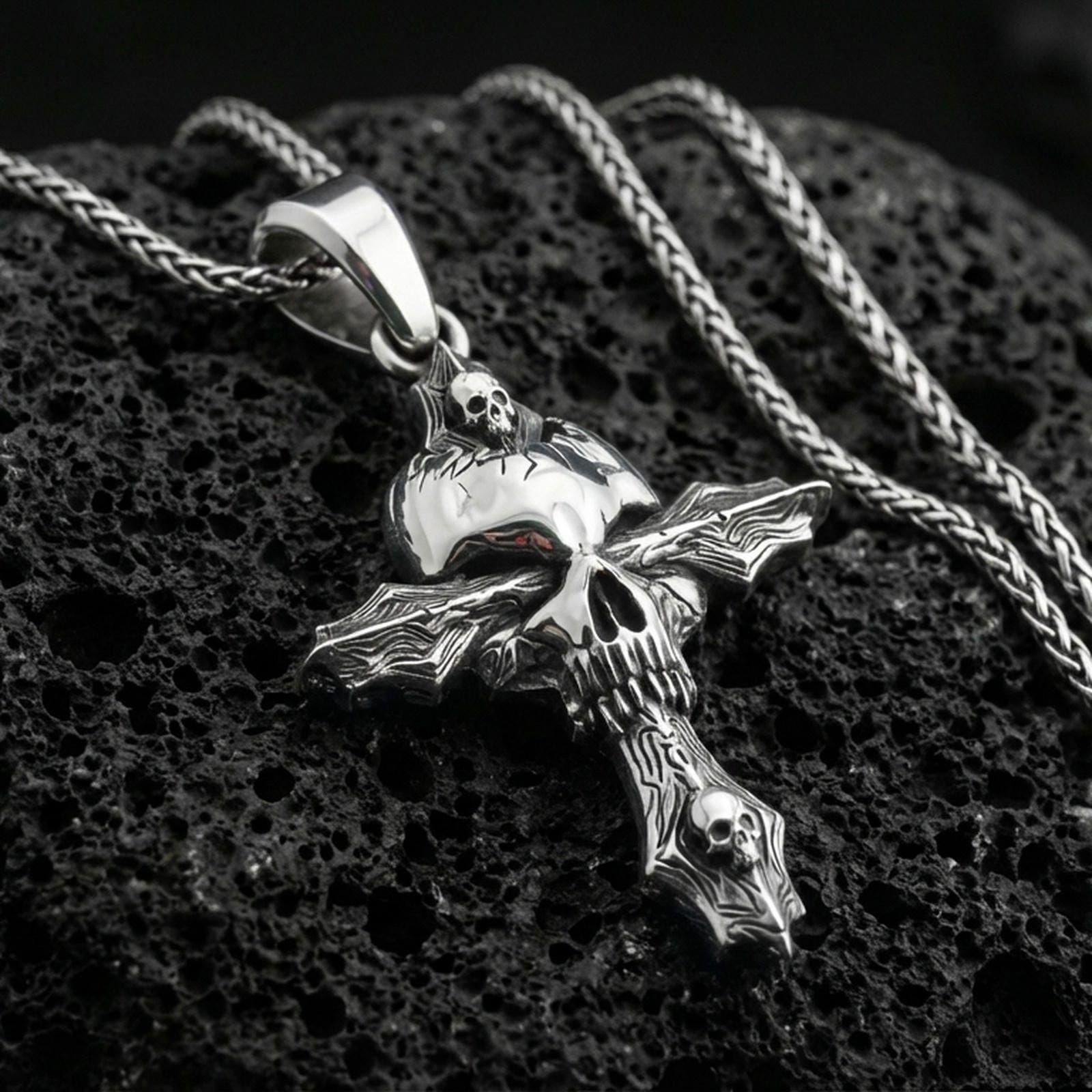 Polished silver gothic cross necklace with central skull motif, carved cross arms, and sculpted ornamental details placed on textured volcanic rock in a premium studio product shot