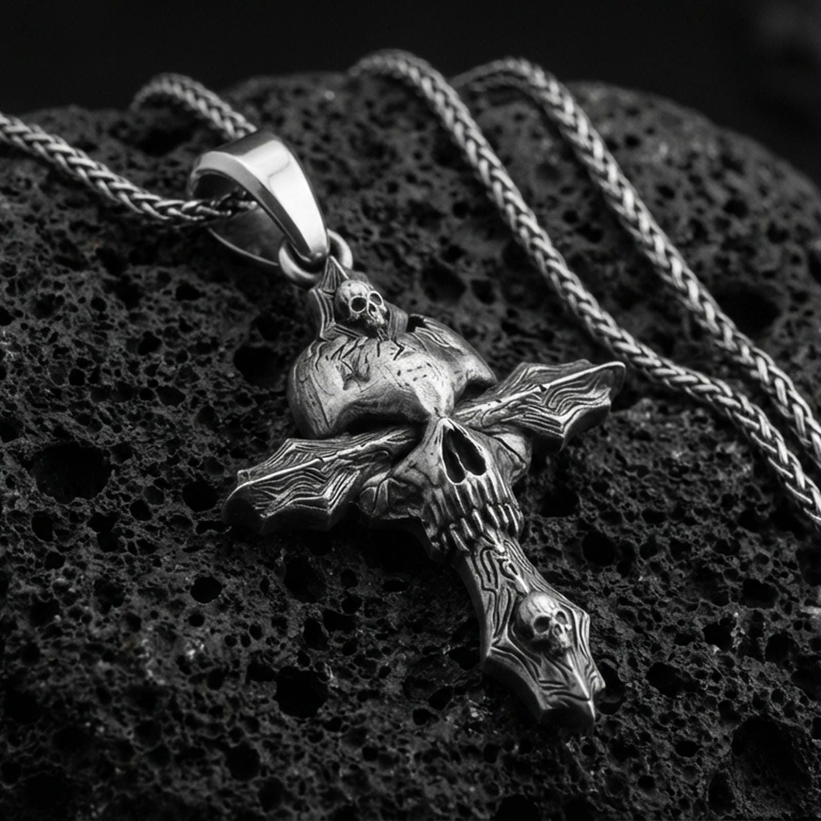 Oxidized gothic skull cross pendant with layered skull details and dark textured relief displayed on dark volcanic rock in a close-up luxury jewelry composition