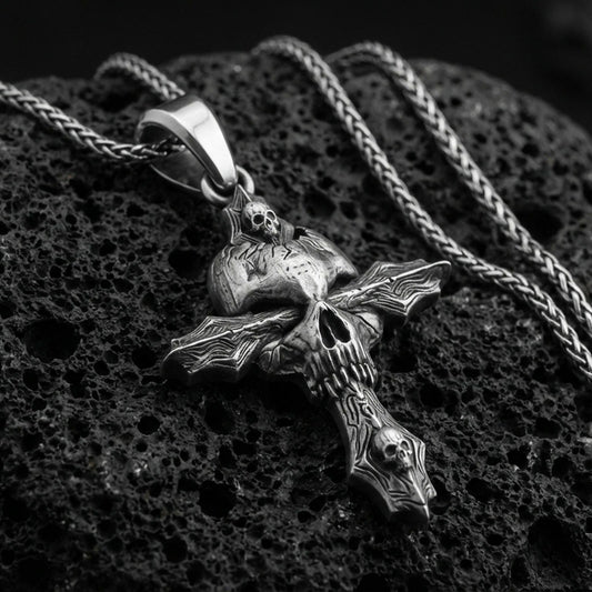 Oxidized gothic skull cross pendant with layered skull details and dark textured relief displayed on dark volcanic rock in a close-up luxury jewelry composition