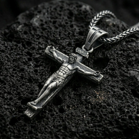 Oxidized crucifix pendant with sculpted Jesus figure, INRI plaque, and wood-textured cross displayed on dark volcanic rock in a close-up luxury jewelry composition