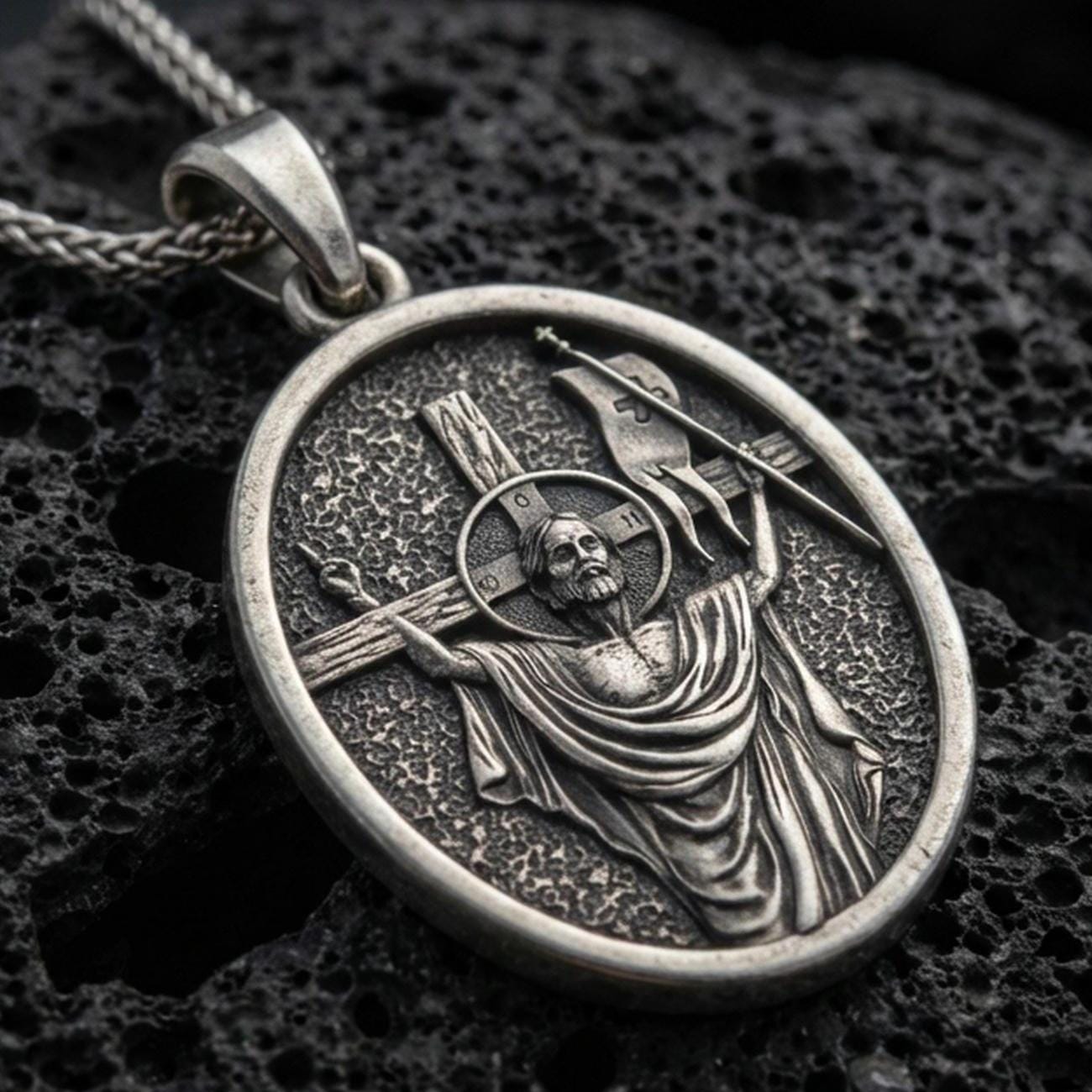 Oxidized resurrection medal pendant with risen Christ, cross staff, and halo detail displayed on dark volcanic rock in a close-up luxury jewelry composition