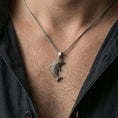 Load image into Gallery viewer, Oxidized dolphin pendant necklace worn on the neck, styled in a dark lifestyle portrait with an open shirt and masculine presentation.
