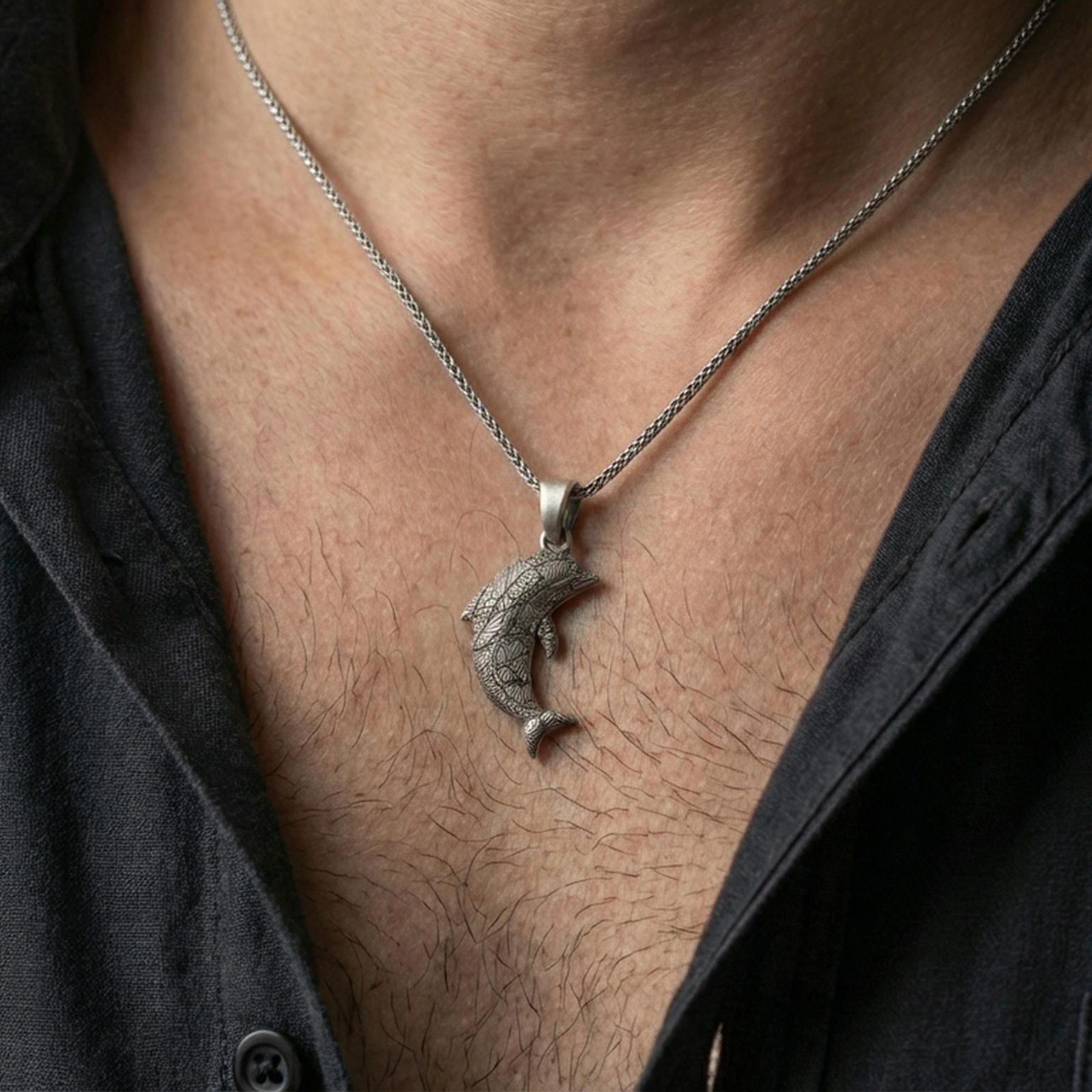 Oxidized dolphin pendant necklace worn on the neck, styled in a dark lifestyle portrait with an open shirt and masculine presentation.