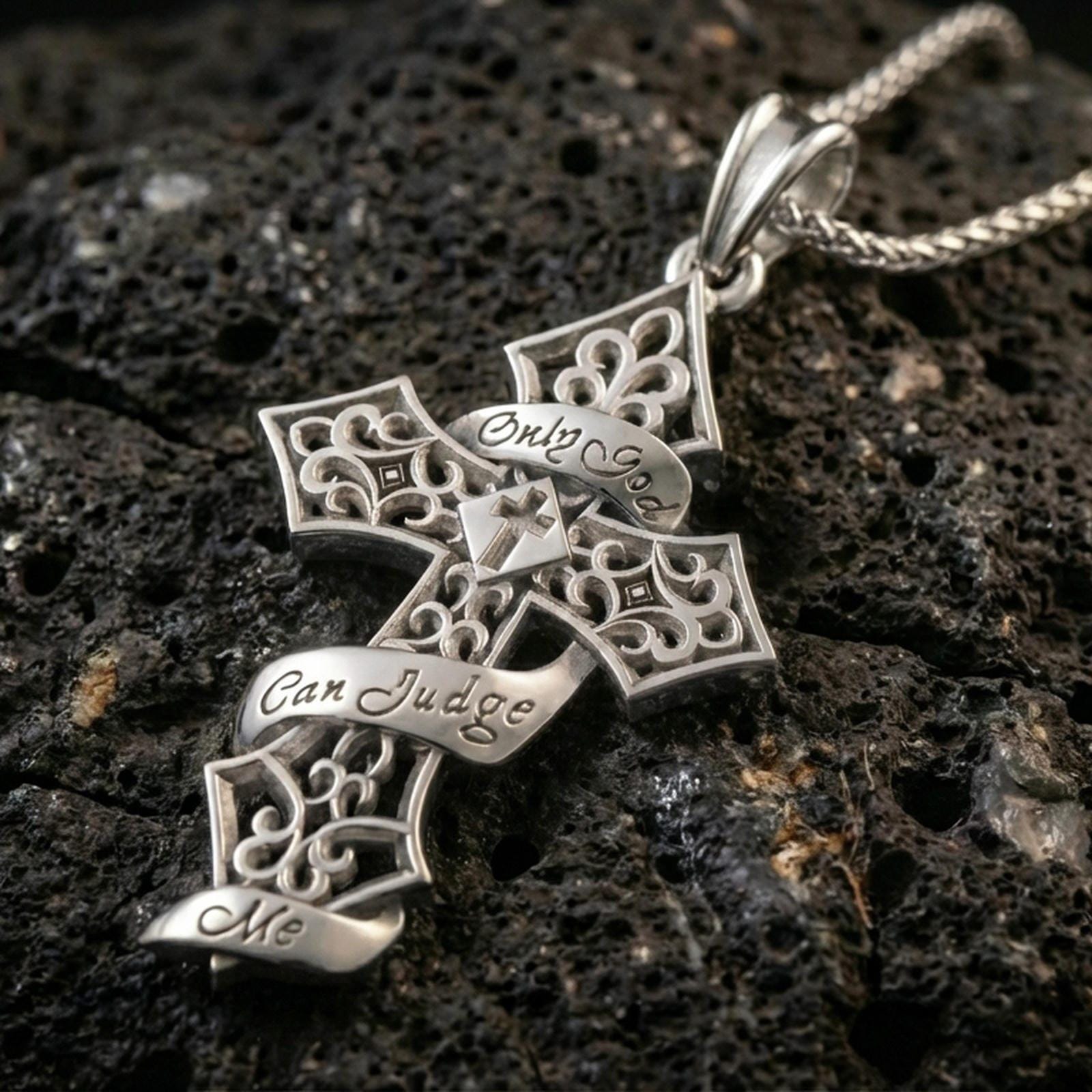 Polished silver Christian cross pendant with openwork gothic pattern, engraved ribbon text, and decorative cross silhouette placed on textured volcanic rock in a premium studio product shot