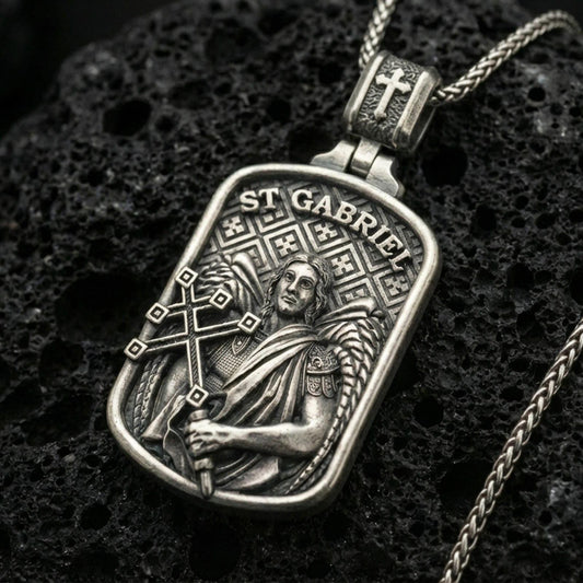 Oxidized Saint Gabriel pendant with angelic figure, wing detail, and ST Gabriel inscription displayed on dark volcanic rock in a close-up luxury jewelry composition